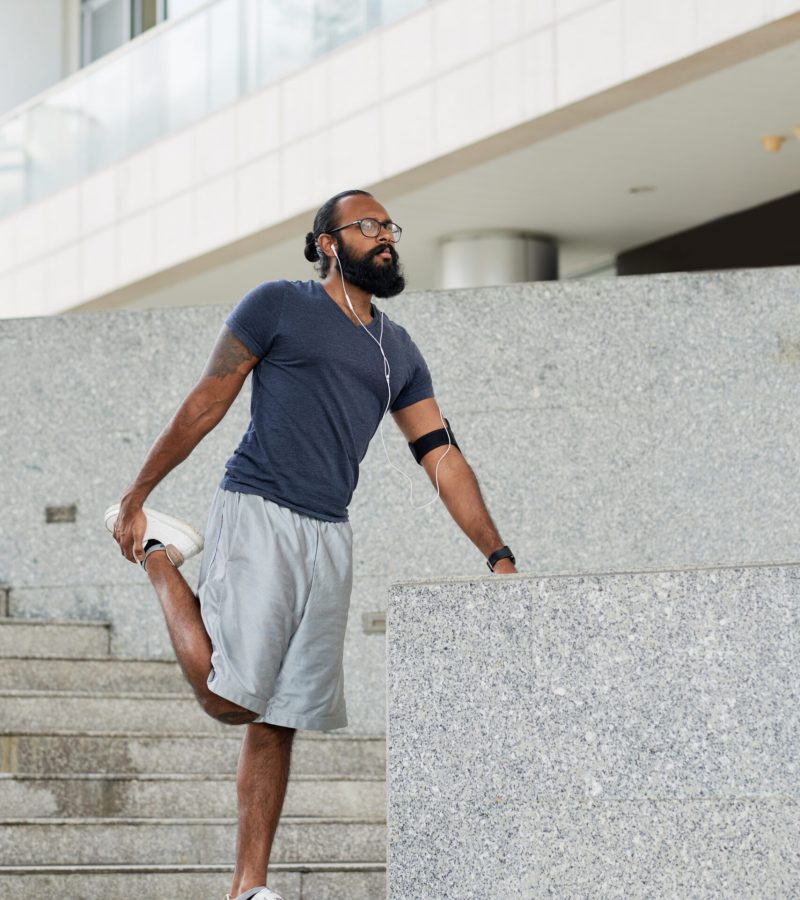 Athletic bearded tattooed Indian man in sportswear listening to music and doing warming up exercises on concrete stairs before running